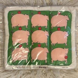 May include: A packaged decorative pillow featuring a repeating pattern of pink pigs with red apples on a green background. The pillow has a white border and is in a clear plastic package.