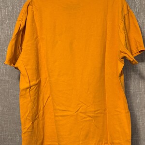Puede incluir: Una camiseta de manga corta en color amarillo mostaza liso con cuello redondo. La prenda se muestra en una percha de pl&aacute;stico transparente, revelando una parte posterior sencilla con un dobladillo est&aacute;ndar y costuras en las mangas.