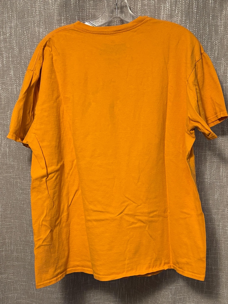Puede incluir: Una camiseta de manga corta en color amarillo mostaza liso con cuello redondo. La prenda se muestra en una percha de pl&aacute;stico transparente, revelando una parte posterior sencilla con un dobladillo est&aacute;ndar y costuras en las mangas.
