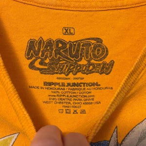 Puede incluir: Una camiseta naranja con una etiqueta de cuello impresa en negro que presenta el logotipo de Naruto Shippuden, talla XL y la marca Ripple Junction. El texto incluye Made in Honduras, 100% algod&oacute;n y la direcci&oacute;n de la empresa en Ohio.