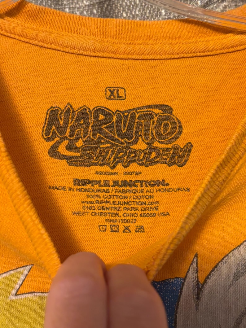 Puede incluir: Una camiseta naranja con una etiqueta de cuello impresa en negro que presenta el logotipo de Naruto Shippuden, talla XL y la marca Ripple Junction. El texto incluye Made in Honduras, 100% algod&oacute;n y la direcci&oacute;n de la empresa en Ohio.