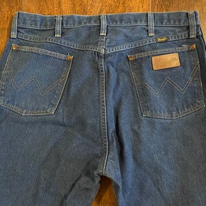 Puede incluir: Un par de pantalones vaqueros de mezclilla azul oscuro con un parche de cuero Wrangler en el bolsillo trasero.