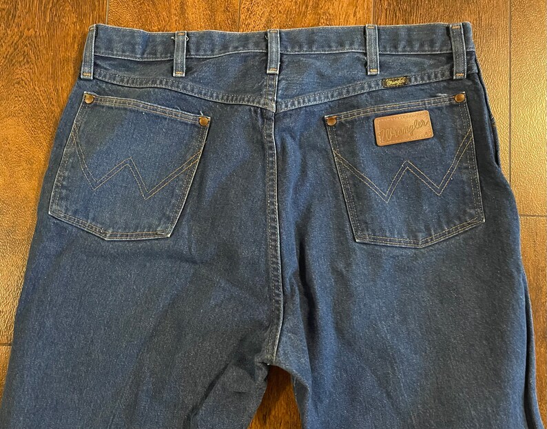 Puede incluir: Un par de pantalones vaqueros de mezclilla azul oscuro con un parche de cuero Wrangler en el bolsillo trasero.