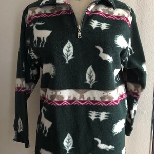 Puede incluir: Un jersey de forro polar verde con estampado de animales blanco y marrón. El jersey tiene una cremallera y un cuello alto.