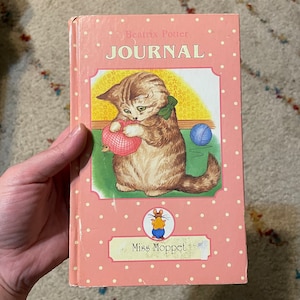 May include: A pink journal with white polka dots and the text "JOURNAL" at the top. The cover features an illustration of a kitten holding a ball of yarn. The journal is titled "Miss Moppet" by Beatrix Potter.