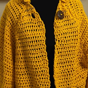 May include: Mustard yellow crochet cardigan with a collared neckline and two wooden buttons. The open weave design of the cardigan is visible, and the garment appears to be handmade. The cardigan is a medium weight and suitable for layering.