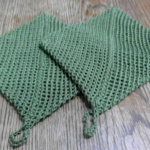 May include: Two square, green crocheted pot holders with a loop for hanging. The pot holders are made of a textured, open weave material. The pot holders are displayed on a wooden surface.