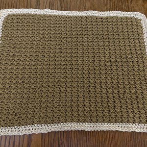 May include: A rectangular, hand-crocheted blanket in a muted olive green color. The blanket has a cream-colored border that frames the textured stitch pattern. The handmade blanket is likely made of soft yarn.
