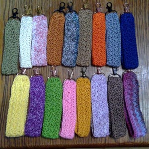 May include: A collection of handmade crochet keychains in various colors, including green, yellow, purple, pink, orange, and blue. Each keychain features a metal clasp for easy attachment. The keychains are arranged on a wooden surface.