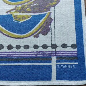 May include: A white linen kitchen towel with a blue border and fruit illustrations. The design includes pears, cherries, and melon slices in purple, yellow, and blue. The towel is signed "T. Tanner".