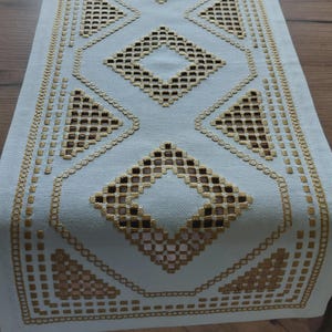 May include: A light blue table runner with a geometric design. The runner features a repeating pattern of diamonds and triangles in a gold color, with openwork details. The runner is made of a soft fabric and is placed on a wooden table.