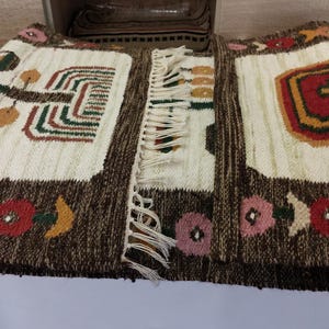 May include: Hand-woven textile with a brown border and cream-colored center panels. The design includes geometric shapes, floral patterns, and fringe. The color palette features brown, red, green, and pink.