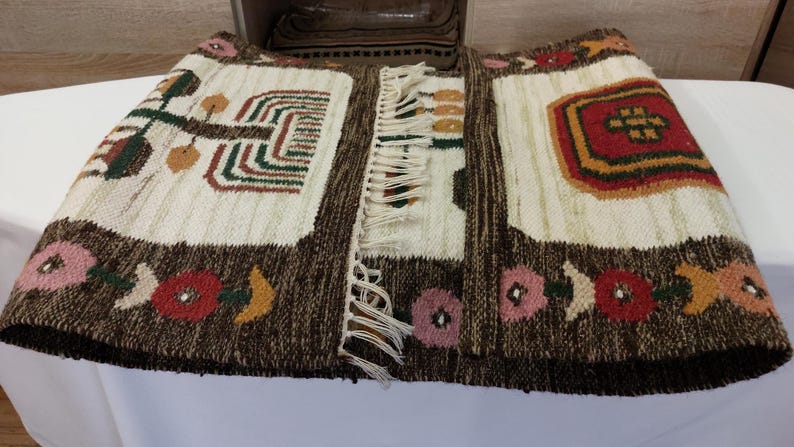 May include: Hand-woven textile with a brown border and cream-colored center panels. The design includes geometric shapes, floral patterns, and fringe. The color palette features brown, red, green, and pink.