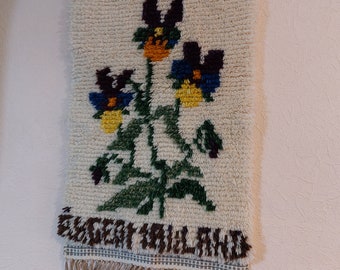 Op de afbeelding: Een witte wandhanger met een bloemenontwerp met drie paarse, gele en blauwe bloemen met groene stelen en bladeren. De wandhanger heeft een bruine franje en de tekst "EUGENIALAND" onderaan.