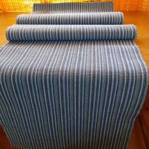 Peut inclure: Un chemin de table bleu et blanc rayé en tissu tissé. Le chemin de table est plié en trois et est posé sur une surface en bois.
