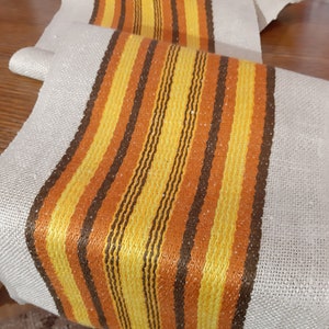 May include: A woven fabric runner with brown, orange, and yellow stripes. The runner is made of a natural linen fabric with a subtle texture.