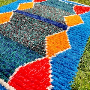 Op de afbeelding: Een kleurrijk, geometrisch tapijt met een blauwe achtergrond en oranje, rode en witte ruitvormen. Het tapijt is gemaakt van een zacht, pluche materiaal.