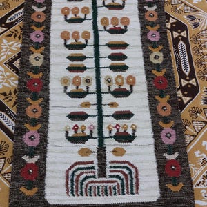 May include: A rectangular, hand-woven rug with a central design of a stylized tree. The rug features a white background with a dark brown border and colorful floral and geometric patterns. The rug has a fringe at the bottom.