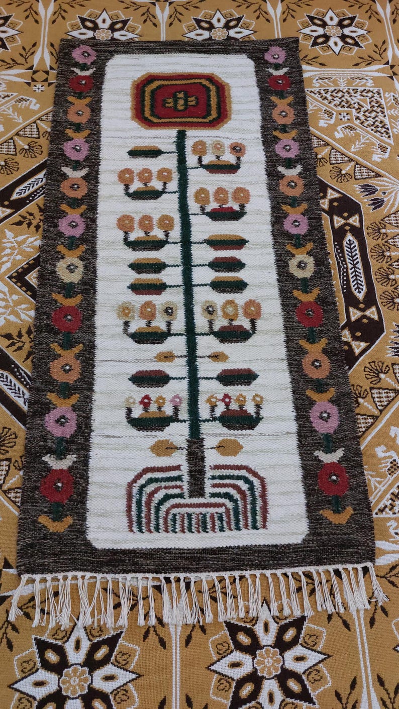 May include: A rectangular, hand-woven rug with a central design of a stylized tree. The rug features a white background with a dark brown border and colorful floral and geometric patterns. The rug has a fringe at the bottom.