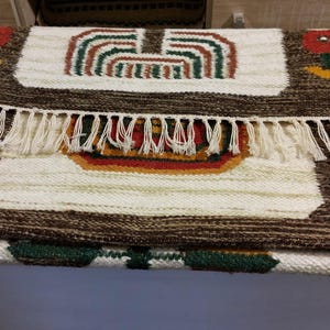 May include: Handmade woven rug with a brown border and cream-colored center. The rug features colorful floral and geometric designs in red, green, and yellow. The rug has a fringe along one edge, adding a decorative touch.