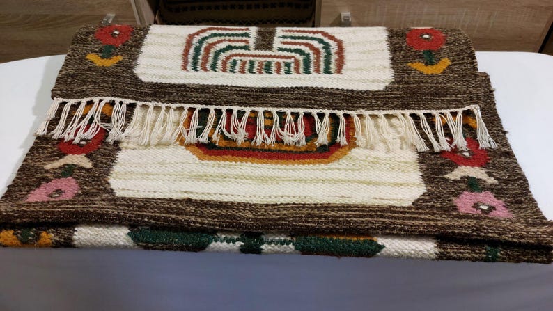 May include: Handmade woven rug with a brown border and cream-colored center. The rug features colorful floral and geometric designs in red, green, and yellow. The rug has a fringe along one edge, adding a decorative touch.