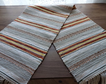 May include: A woven table runner with a striped pattern in shades of white, beige, orange, and yellow. The runner has a fringed edge.
