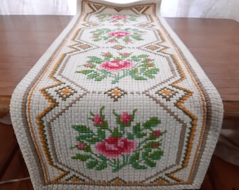 May include: White table runner with a floral design featuring pink roses and green leaves. The runner has a brown and yellow border.