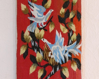 Op de afbeelding: Een rood wandkleed met een geweven ontwerp van twee blauwe en witte vogels die op een tak zitten met zwarte en groene bladeren. De achtergrond is effen rood.