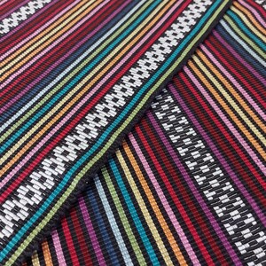 May include: Multicolored striped fabric with a white and black geometric pattern. The fabric is woven with a variety of colors, including red, orange, yellow, green, blue, purple, and pink.
