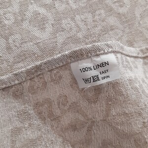 May include: A white fabric label with the text "100% LINEN" and "EASY SPIN" in black text. The label is attached to a beige fabric with a floral pattern.