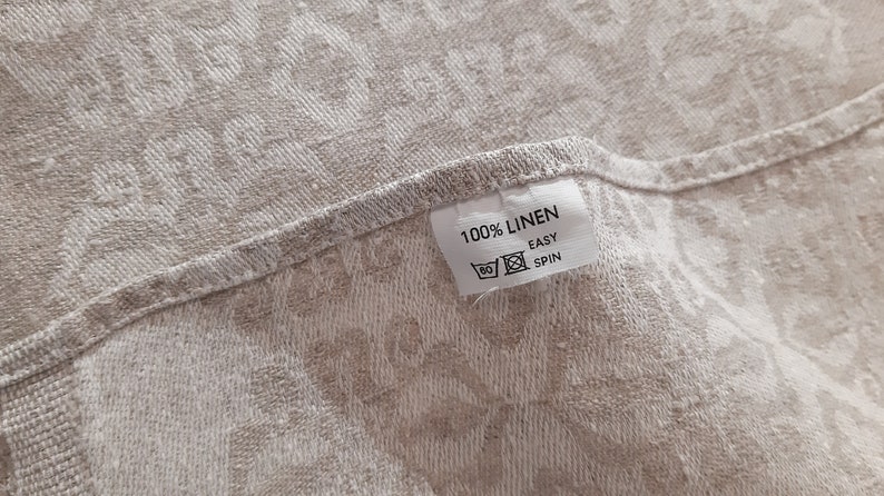 May include: A white fabric label with the text "100% LINEN" and "EASY SPIN" in black text. The label is attached to a beige fabric with a floral pattern.