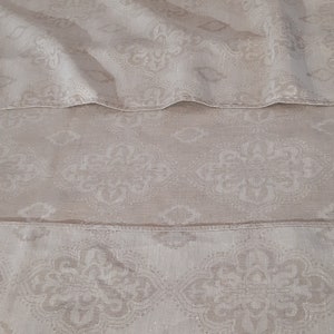 May include: A close-up of a beige fabric with a subtle, repeating pattern of white floral designs. The fabric is folded in three layers, creating a layered effect.