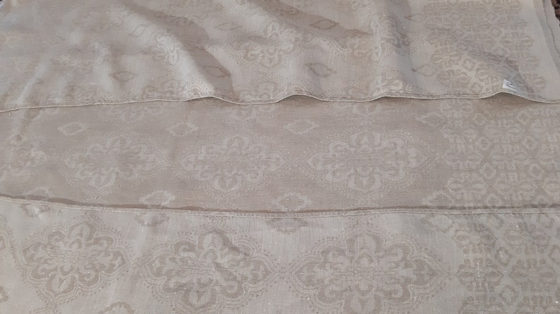 May include: A close-up of a beige fabric with a subtle, repeating pattern of white floral designs. The fabric is folded in three layers, creating a layered effect.