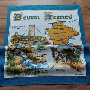 May include: A blue and white tea towel with a map of Devon, England, and images of the Tamar Bridge, Babbacombe Bay, and Clovelly. The text "Devon Scenes" is printed in a decorative font at the top of the towel. The text "Made in Britain" is printed at the bottom of the towel.