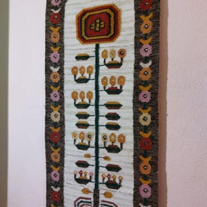 May include: A woven wall hanging with a central floral design in red, yellow, and brown. The design is on a cream-colored background with a dark brown border. The border features repeating floral patterns in various colors.