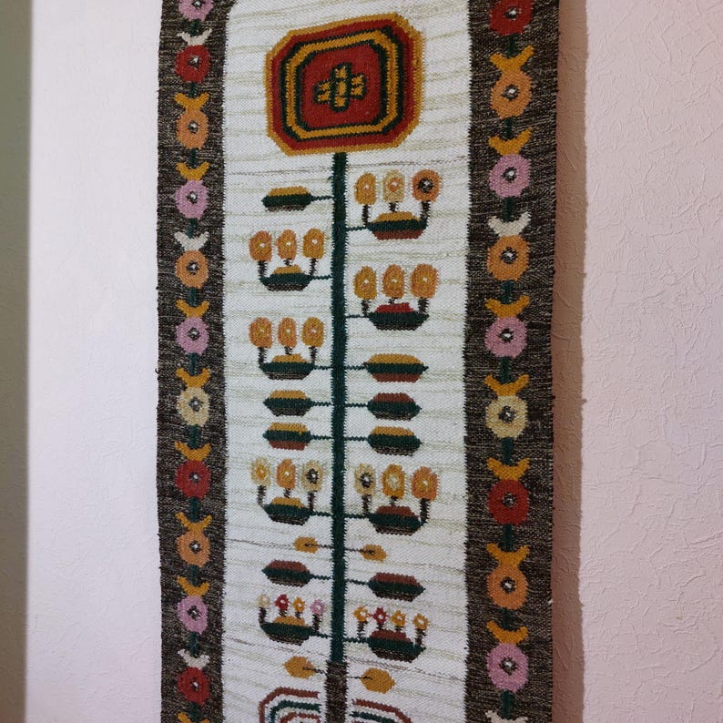 May include: A woven wall hanging with a central floral design in red, yellow, and brown. The design is on a cream-colored background with a dark brown border. The border features repeating floral patterns in various colors.