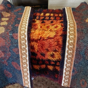 Op de afbeelding: Twee kleurrijke tapijten met franjes aan de randen. Een tapijt is een donkerblauw en oranje geometrisch patroon met een bruine kettingrand. Het andere tapijt is een ruig oranje en bruin tapijt met een geometrisch patroon.