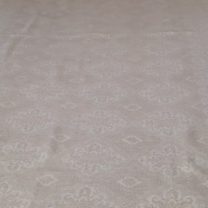 May include: A close-up of a white fabric with a subtle, embossed pattern of repeating floral designs. The fabric has a slightly textured surface.