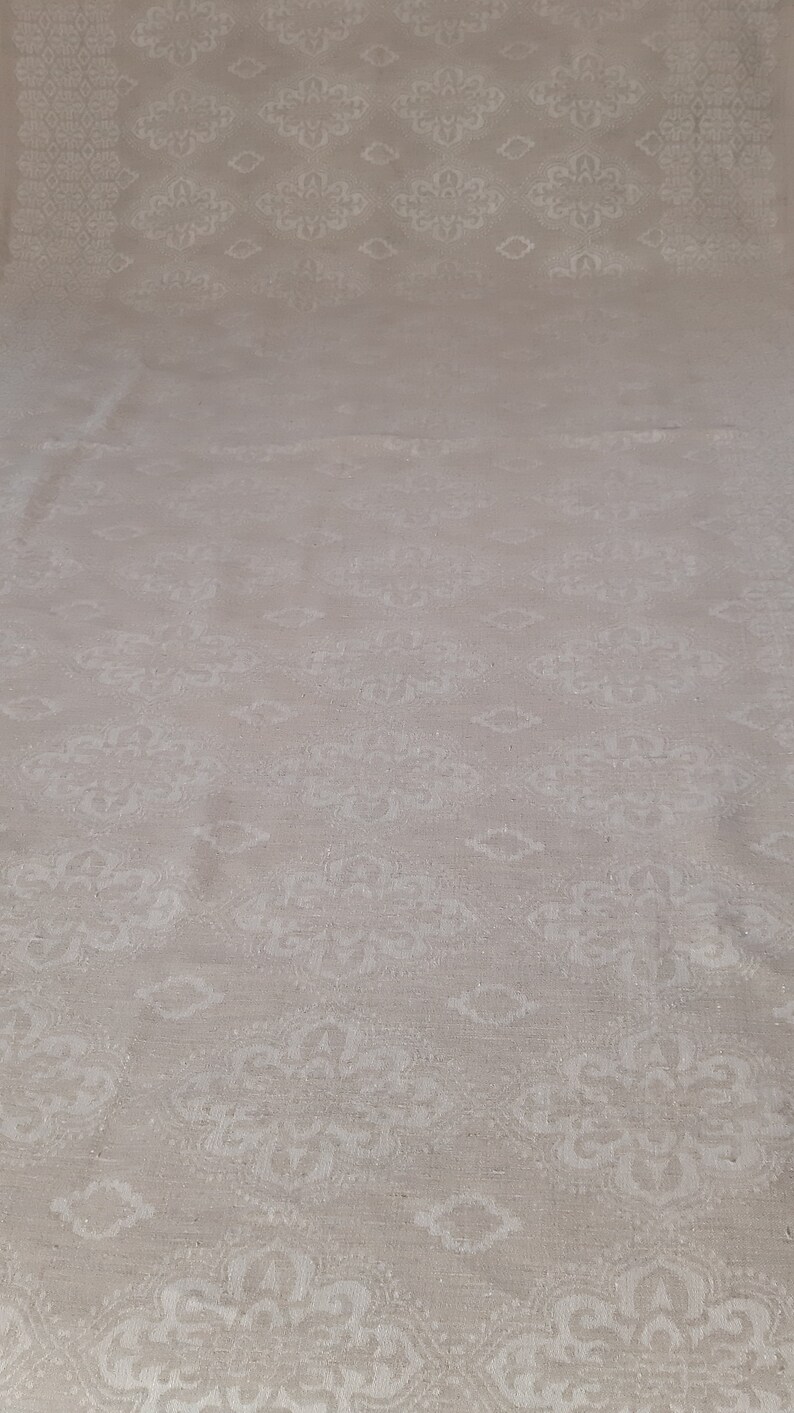 May include: A close-up of a white fabric with a subtle, embossed pattern of repeating floral designs. The fabric has a slightly textured surface.
