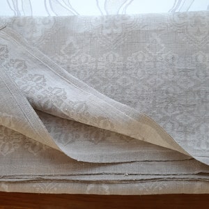 May include: A folded white linen tablecloth with a damask pattern. The tablecloth is folded in thirds and has a slightly textured surface.