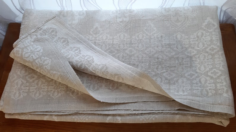 May include: A folded white linen tablecloth with a damask pattern. The tablecloth is folded in thirds and has a slightly textured surface.
