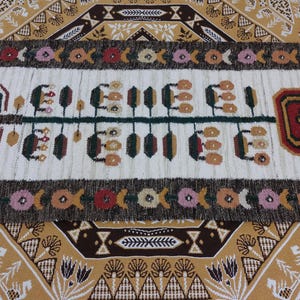 May include: Rectangular, hand-woven rug with a cream-colored central field featuring a stylized design in green, brown, and tan. The rug is bordered by a dark brown band with floral and geometric patterns. Fringes are visible on the short ends.