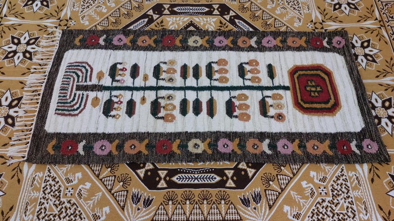 May include: Rectangular, hand-woven rug with a cream-colored central field featuring a stylized design in green, brown, and tan. The rug is bordered by a dark brown band with floral and geometric patterns. Fringes are visible on the short ends.