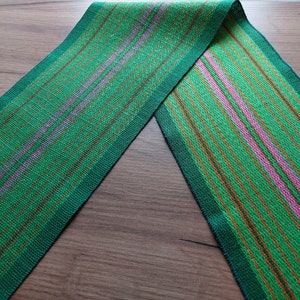 Op de afbeelding: Een geweven lint in groen, oranje en roze met strepen. Het lint is doormidden gevouwen en de twee uiteinden wijzen naar elkaar.