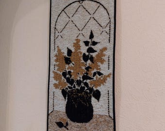 May include: A beige and brown tapestry wall hanging with a black floral design in a vase. The tapestry has a black border and a window-like design with a grid pattern.