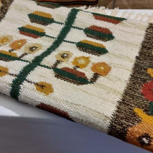 May include: Hand-woven rug with a cream-colored background and a central design of a green stem with stylized flowers. The border is brown with floral motifs in red, yellow, and orange. The rug has a fringed edge.
