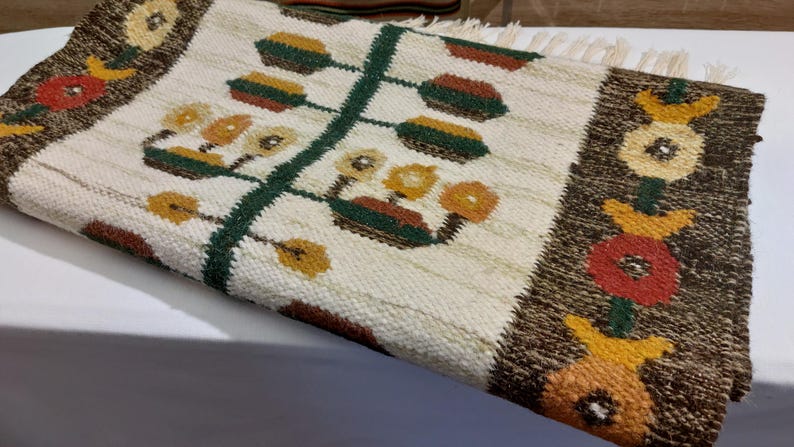 May include: Hand-woven rug with a cream-colored background and a central design of a green stem with stylized flowers. The border is brown with floral motifs in red, yellow, and orange. The rug has a fringed edge.