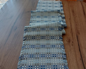 May include: A blue and white woven table runner with a fringed edge. The runner has a geometric pattern and is made of a natural fiber.