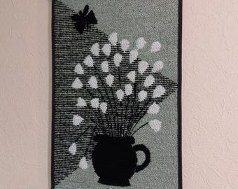 May include: A black and white tapestry with a floral design. The flowers are white and the vase is black. The background is a light green color.