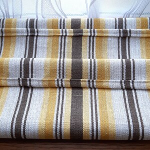 May include: A striped table runner with brown and yellow stripes on a white background. The runner is folded in thirds and is lying on a wooden surface.